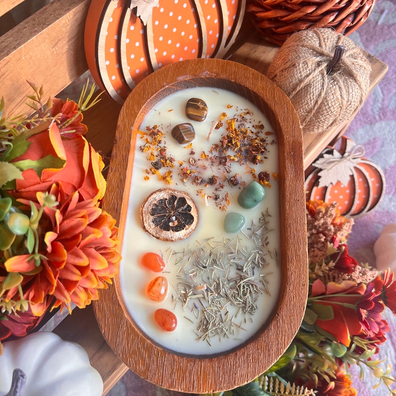 May include: A wooden bowl candle with a white wax base and various natural elements embedded in the wax, including dried herbs, crystals, and a dried citrus slice. The bowl is surrounded by fall-themed decorations.