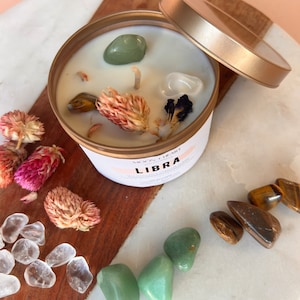 May include: A candle in a gold tin with a white lid. The candle is decorated with dried flowers, crystals, and the text "LIBRA" in pink lettering. The candle is surrounded by various crystals, including clear quartz, green aventurine, and tiger's eye.