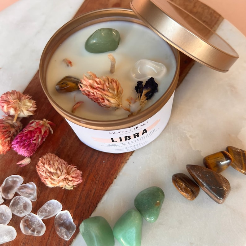 May include: A candle in a gold tin with a white lid. The candle is decorated with dried flowers, crystals, and the text "LIBRA" in pink lettering. The candle is surrounded by various crystals, including clear quartz, green aventurine, and tiger's eye.