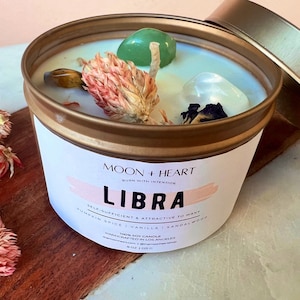 May include: A gold candle tin with a white label that reads "MOON + HEART BURN WITH INTENTION LIBRA SELF-SUFFICIENT & ATTRACTIVE TO MANY PUMPKIN SPICE | VANILLA | SANDALWOOD 100% SOY CANDLE HANDCRAFTED IN LOS ANGELES moonheartcandle.com | @moonheartcandle 8 OZ | 225 G". The candle is topped with dried flowers, a green crystal, and a clear crystal.