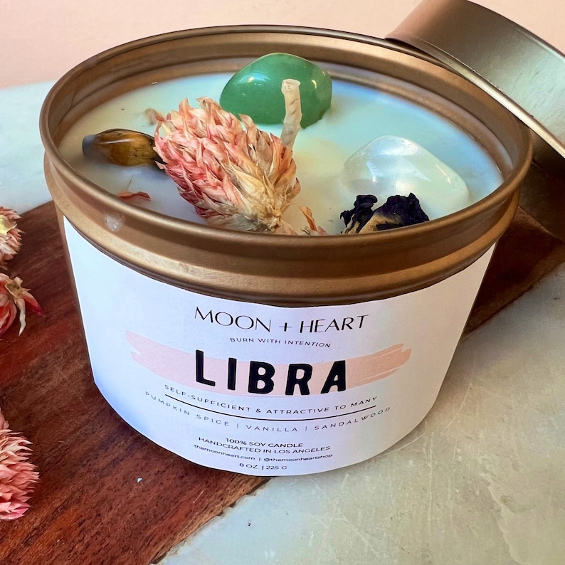 May include: A gold candle tin with a white label that reads "MOON + HEART BURN WITH INTENTION LIBRA SELF-SUFFICIENT & ATTRACTIVE TO MANY PUMPKIN SPICE | VANILLA | SANDALWOOD 100% SOY CANDLE HANDCRAFTED IN LOS ANGELES moonheartcandle.com | @moonheartcandle 8 OZ | 225 G". The candle is topped with dried flowers, a green crystal, and a clear crystal.