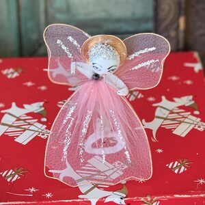 May include: A vintage Christmas tree topper angel ornament with a pink tulle skirt, gold halo, and white face with painted features.