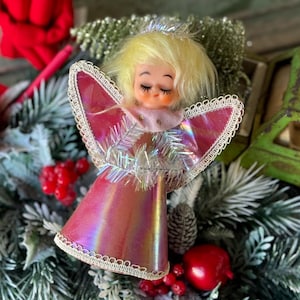 May include: A vintage Christmas angel ornament with a pink iridescent dress and wings, trimmed with white lace. The angel has closed eyes, blonde hair, and a silver halo. The ornament is surrounded by faux evergreen branches and red berries.
