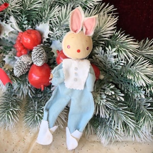 May include: A blue and white fabric bunny doll with pink ears, sitting in a snowy evergreen wreath. The doll has a white bib with three buttons and a round, painted face.