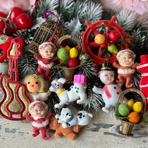 May include: A collection of vintage Christmas ornaments, including a red guitar, a snowman, and figures of children in red and white outfits. There are also miniature baskets with fruit and Dalmatian dog figurines. The ornaments are arranged on a surface with faux evergreen branches.