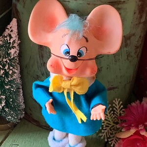 May include: A vintage toy mouse figurine with a light pink face, large ears, and blue eyes. The mouse wears a blue outfit with a yellow bow tie and white trim. A small Christmas tree is in the background.