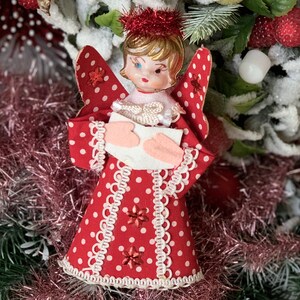 May include: A red and white polka dot angel ornament with a white lace trim and a pink heart in its hands. The angel has blonde hair and a red and white hat. The ornament is hanging on a Christmas tree.