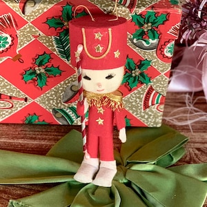 May include: A vintage Christmas ornament in the shape of a toy soldier. The red felt figure has a matching hat with gold stars, a gold collar, and holds a candy cane. It is set against a red and green patterned gift box and a green bow.