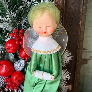 May include: A vintage angel figurine with a serene expression, green hair, and a green satin dress. The angel has a white collar with gold trim and delicate wings. Red berries and faux pine branches are in the background.