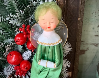 May include: A vintage angel figurine with a serene expression, green hair, and a green satin dress. The angel has a white collar with gold trim and delicate wings. Red berries and faux pine branches are in the background.
