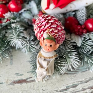 May include: A vintage Christmas ornament in the shape of an elf. The elf has a painted face, a green hat, and a white outfit with gold trim. The ornament is hanging in front of a Christmas tree with red berries and pinecones.