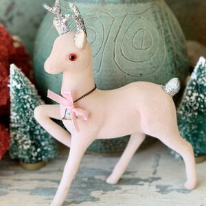 May include: A pink flocked reindeer figurine with silver glitter antlers and a pink ribbon around its neck. The reindeer is wearing a silver bell with a blue and white design.