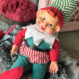 May include: A vintage Christmas elf doll with a smiling face, wearing a red and green outfit. The elf has a white collar, a striped red and white skirt, and a green and white striped hat. The doll is surrounded by Christmas decorations.