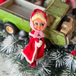 May include: A vintage Christmas ornament featuring a doll with blonde hair, blue eyes, and a red coat with white trim. The doll is standing in front of a green toy truck. The ornament is placed on a bed of faux evergreen branches.