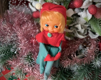 May include: A vintage Christmas elf doll with blonde hair, blue eyes, and freckles. The elf wears a red hat with a white pom-pom, a red collar, and a teal outfit. The doll is surrounded by pink tinsel and Christmas decorations.