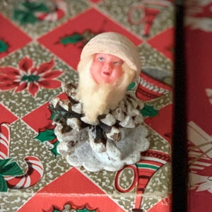 May include: A vintage Christmas ornament featuring a Santa Claus figure with a white beard and hat, crafted from a pine cone. The ornament has a painted face and is set against a red and gold patterned background with holiday motifs.