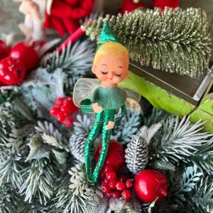 May include: A vintage Christmas elf figurine with a green outfit and a pointed hat, set amidst festive decorations. The elf has a serene expression and is surrounded by red ornaments, pine needles, and a glittery green tree.