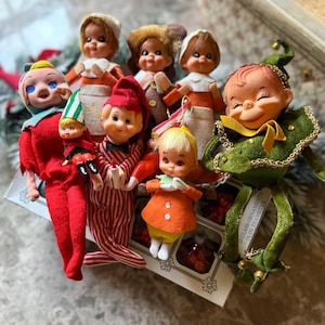May include: A collection of vintage-style elf ornaments with felt clothing in red, green, orange, and white stripes. The figures feature painted faces and are positioned atop a box labeled 12 glass ornaments 2 in. in diameter.
