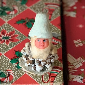 May include: A vintage Christmas gnome figurine with a light blue hat and a pink face. The gnome's beard is made of white material, and its body is made of pinecones. The figurine is placed on a red and green patterned surface.