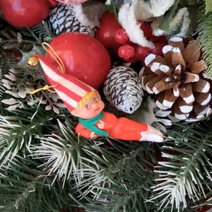 May include: A small red and white elf figurine with a green scarf and red pants is nestled in a green and white Christmas wreath. The wreath is decorated with pine cones, berries, and snow-covered branches.
