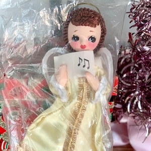 May include: A vintage Christmas angel ornament in a plastic bag. The angel has a yellow dress, white wings, and a halo. She holds a small card with musical notes. The angel has rosy cheeks and brown hair. A pink tinsel Christmas tree is in the background.