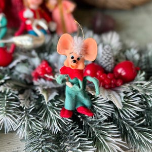 May include: A vintage Christmas ornament in the shape of a mouse. The mouse has large pink ears, a red collar, and a green outfit with red shoes. The ornament is set against a backdrop of frosted evergreen branches and red berries.