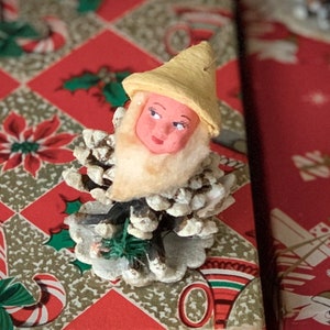 May include: A vintage Christmas ornament featuring a gnome figure with a painted face, yellow hat, and white beard, attached to a pinecone base. The ornament is set against a festive red and gold patterned background.