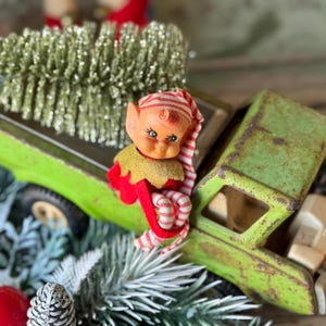 May include: A vintage green toy truck with a gold glittery Christmas tree in the bed. A small elf doll with a red and white striped hat and outfit is perched on the truck. The truck has a weathered appearance.