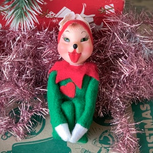May include: Vintage Christmas elf figurine with a painted face, red hat and collar, and green felt body. The elf has a red heart on its chest and white gloves. The background includes pink tinsel and a Christmas-themed box.