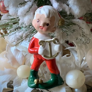 May include: A ceramic Christmas elf figurine, dressed in a red outfit with green shoes, holding a white harp. The elf has white hair and a rosy complexion. The figurine is surrounded by white decorations and ornaments.