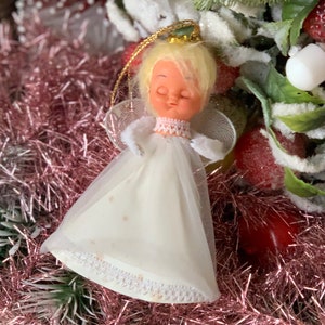 May include: A vintage Christmas angel ornament with a blonde head, white dress, and delicate wings. The angel has closed eyes and a gold halo. The ornament is set against a backdrop of pink tinsel and festive decorations.