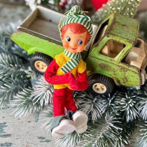 May include: A vintage Christmas ornament of an elf wearing a green and white striped hat and scarf, a yellow vest, red pants and mittens. The elf is standing in front of a green toy truck. The ornament is on a bed of faux evergreen branches.