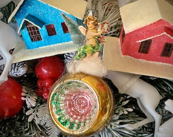 May include: A vintage Christmas ornament featuring a gold-colored glass ball with a rose design, surrounded by a wire mesh. An angel figurine with green and white clothing is perched above the ball. Miniature blue and red houses with white snow-covered roofs are also visible.