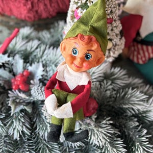 May include: A vintage Christmas elf doll with a green hat, red and white outfit, and green pants. The elf has a smiling face with blue eyes and is sitting on a bed of frosted greenery. A festive holiday decoration.