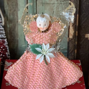May include: A vintage Christmas tree topper in the shape of an angel. The angel has a porcelain face, a pink dress with gold trim, and silver and gold mesh wings. A white poinsettia and green leaf adorn the front of the dress.