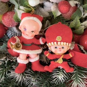 May include: Two vintage Christmas ornaments. One is a Santa Claus figure in a red suit with white trim, holding a gold horn. The other is a red soldier figure with a black belt and gold accents. Both are set against a backdrop of festive greenery.