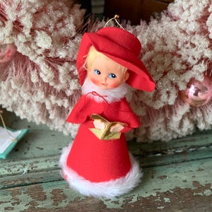 May include: A vintage Christmas ornament featuring a girl wearing a red dress and hat, holding a book. The ornament is made of felt and has a white fur trim.
