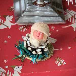 May include: A vintage Christmas ornament featuring a Santa Claus figure. The Santa has a pink face, white beard, and a cream-colored hat. He is surrounded by a pine cone body and blue tinsel. The ornament sits on a red surface with a Christmas-themed pattern.