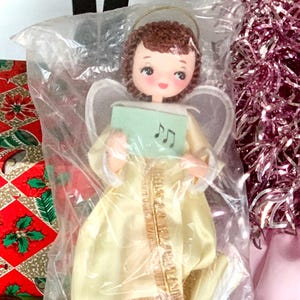 May include: A vintage Christmas angel ornament in a plastic bag. The angel has a yellow gown, brown hair, and holds a green card with musical notes. A red and gold patterned gift box and a pink tinsel tree are also visible.
