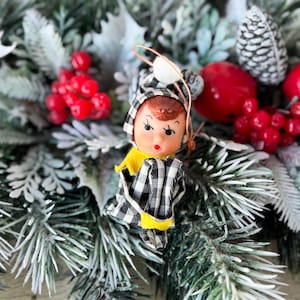 May include: A vintage Christmas ornament featuring a doll dressed in a black and white checkered outfit with a yellow collar. The ornament is set against a backdrop of faux evergreen branches, red berries, and pinecones.