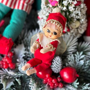 May include: A vintage Christmas elf doll with a red outfit, hat, and gold trim. The doll has a painted face and is surrounded by festive decorations, including pine cones and berries. The elf is a classic holiday decoration.