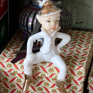 May include: A ceramic elf figurine with a white outfit, gold boots, and a gold-tipped hat. The elf is posed with one arm on its hip. The figurine is sitting on a box with a red and green floral pattern.