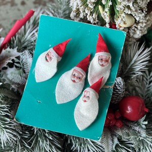 May include: A vintage Christmas card featuring four felt Santa Claus ornaments with red hats and white beards. The card is teal and surrounded by festive greenery and red berries, evoking a nostalgic holiday theme.