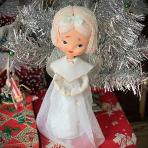 May include: A vintage Christmas angel doll with a white dress, gold accents, and a light green bow. The doll has a painted face and is positioned in front of a silver tinsel Christmas tree. The angel is a festive holiday decoration.