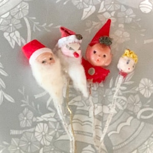 May include: A collection of vintage Christmas ornaments featuring Santa Claus, an elf, and a boy. Each ornament has a red hat and a white beard or hair. The ornaments are attached to clear stems, set against a floral background.