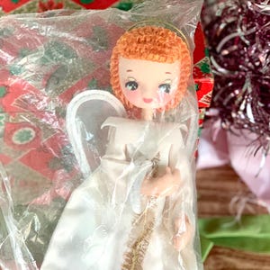 May include: A vintage Christmas angel doll with orange hair, a gold halo, and white gown, packaged in clear plastic. The doll has a sweet face with blue eyes and is surrounded by festive holiday decorations.