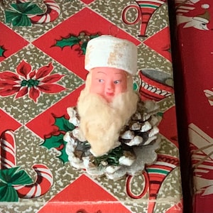 May include: Vintage Christmas ornament featuring Santa Claus with a white beard and hat, attached to a pinecone base. The ornament is set against a red and gold patterned background with candy canes and poinsettias.