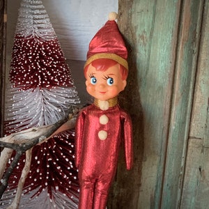 May include: A vintage Christmas elf doll with a red metallic jumpsuit and matching hat. The doll has a painted face with blue eyes and a gold collar. A small white and red Christmas tree is in the background.