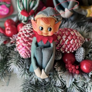 May include: A vintage Christmas elf ornament with a gray hat, red collar, and green outfit. The elf has rosy cheeks and is surrounded by festive decorations, including red apples, pinecones, and evergreen branches. A charming holiday decoration.