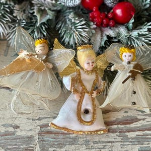 May include: Three vintage angel Christmas ornaments. Each angel has a white dress, gold wings, and a painted face. One angel holds a musical instrument. The ornaments are set against a backdrop of evergreen branches and red berries.